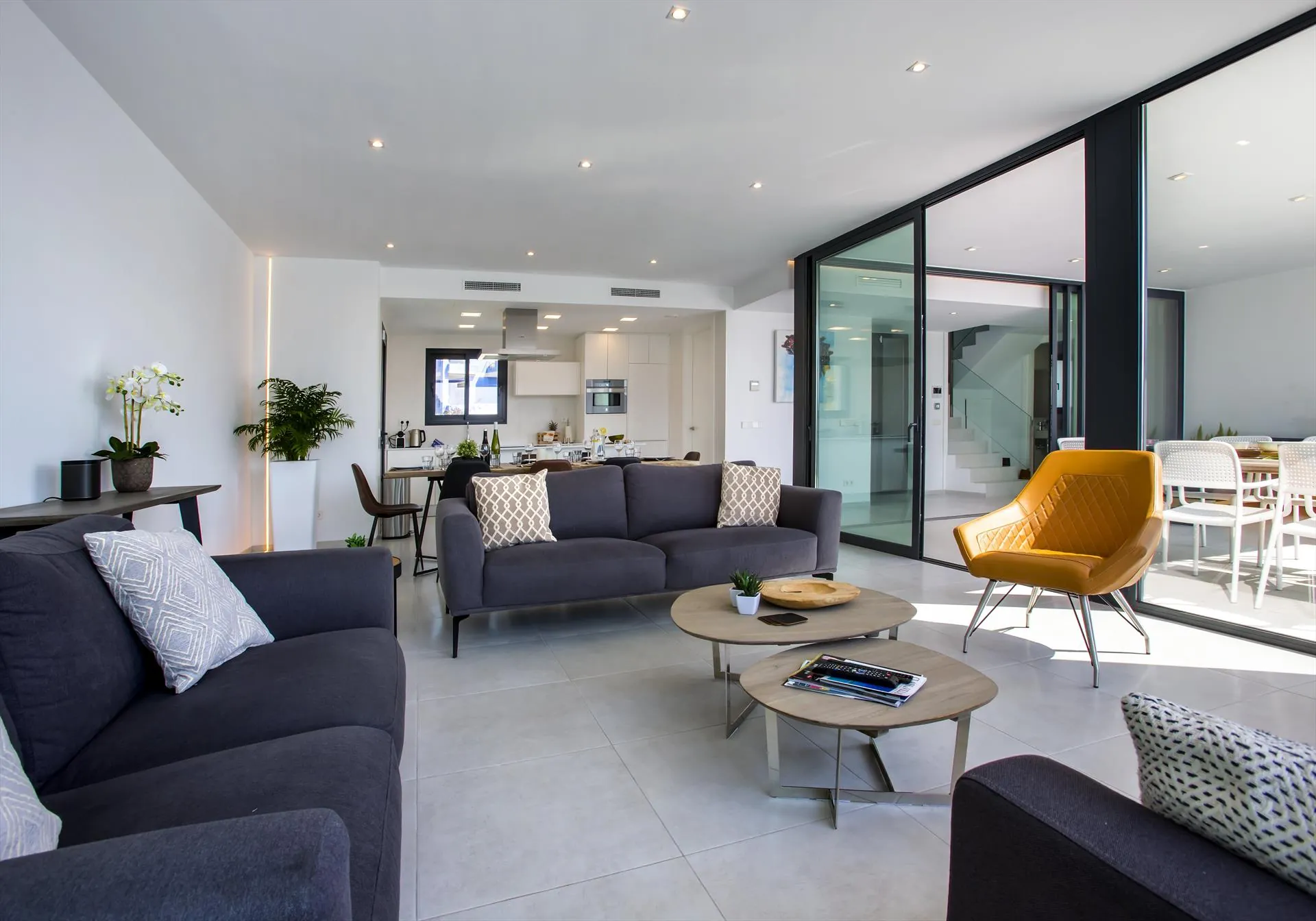 De woonkamer met een diepe grijze bank, een okergele fauteuil en twee ronde tafels.
