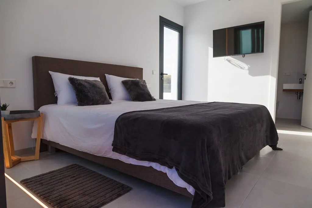 Een slaapkamer met een donkere sprei, een kleed naast het bed en een televisie aan de wand.