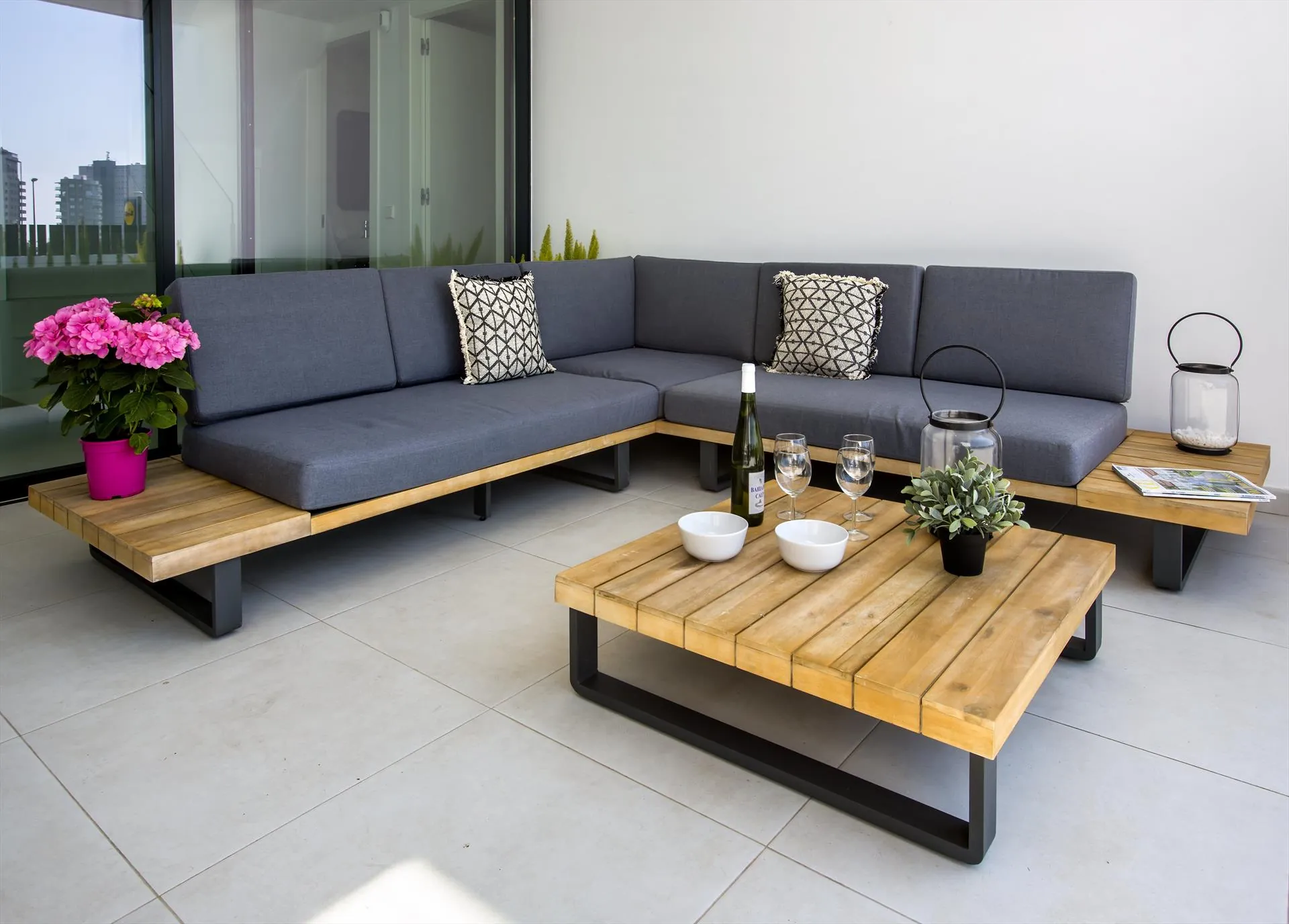 Het overdekte terras met een donkergrijze hoekbank om twee lage houten tafels, bloemen en lantaarns.